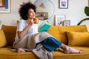 A woman reading a book at home, drinking coffee sitting on the couch.